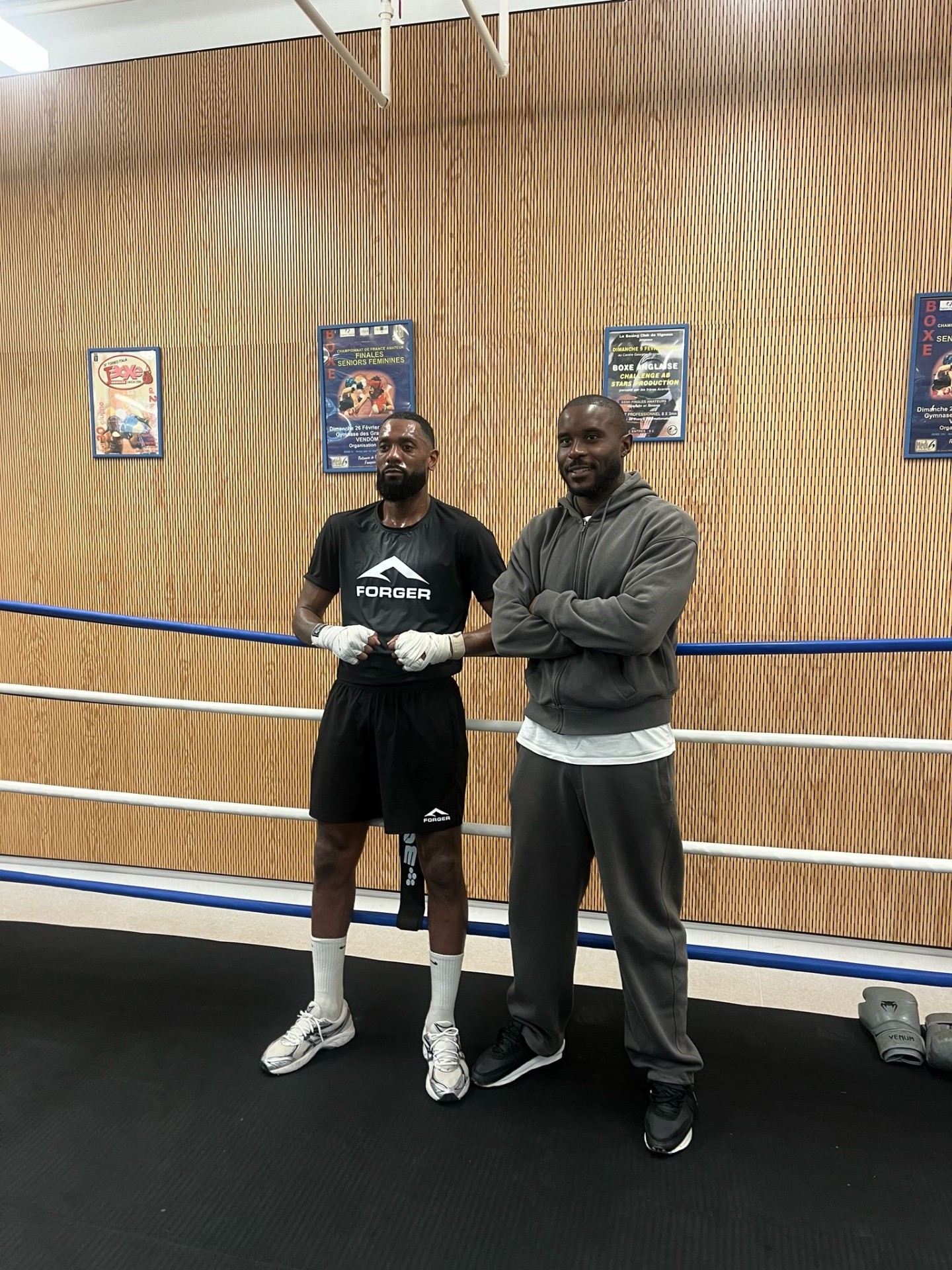 Deux membres de la communauté FORGER posent dans un ring de boxe — t-shirt et short FORGER noirs, mains bandées, esprit de famille et discipline partagée